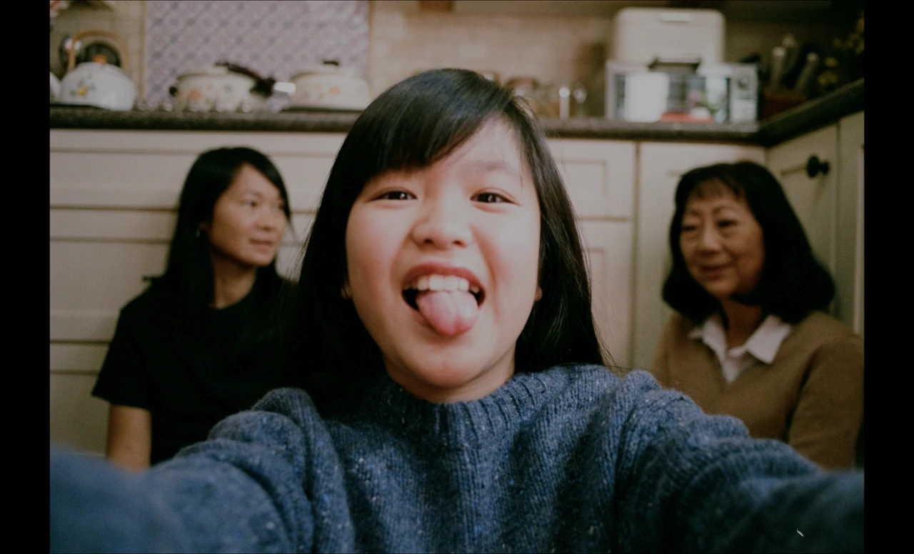 A child smiles and sticks out her tongue toward the camera, with two women seated behind her in a kitchen.