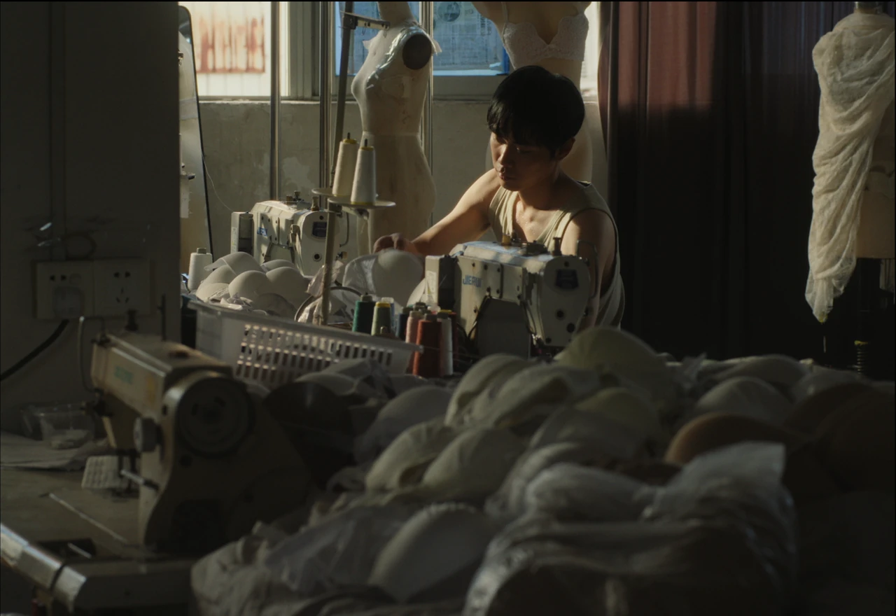 A young person working at a sewing machine among stacks of lingerie in a factory.